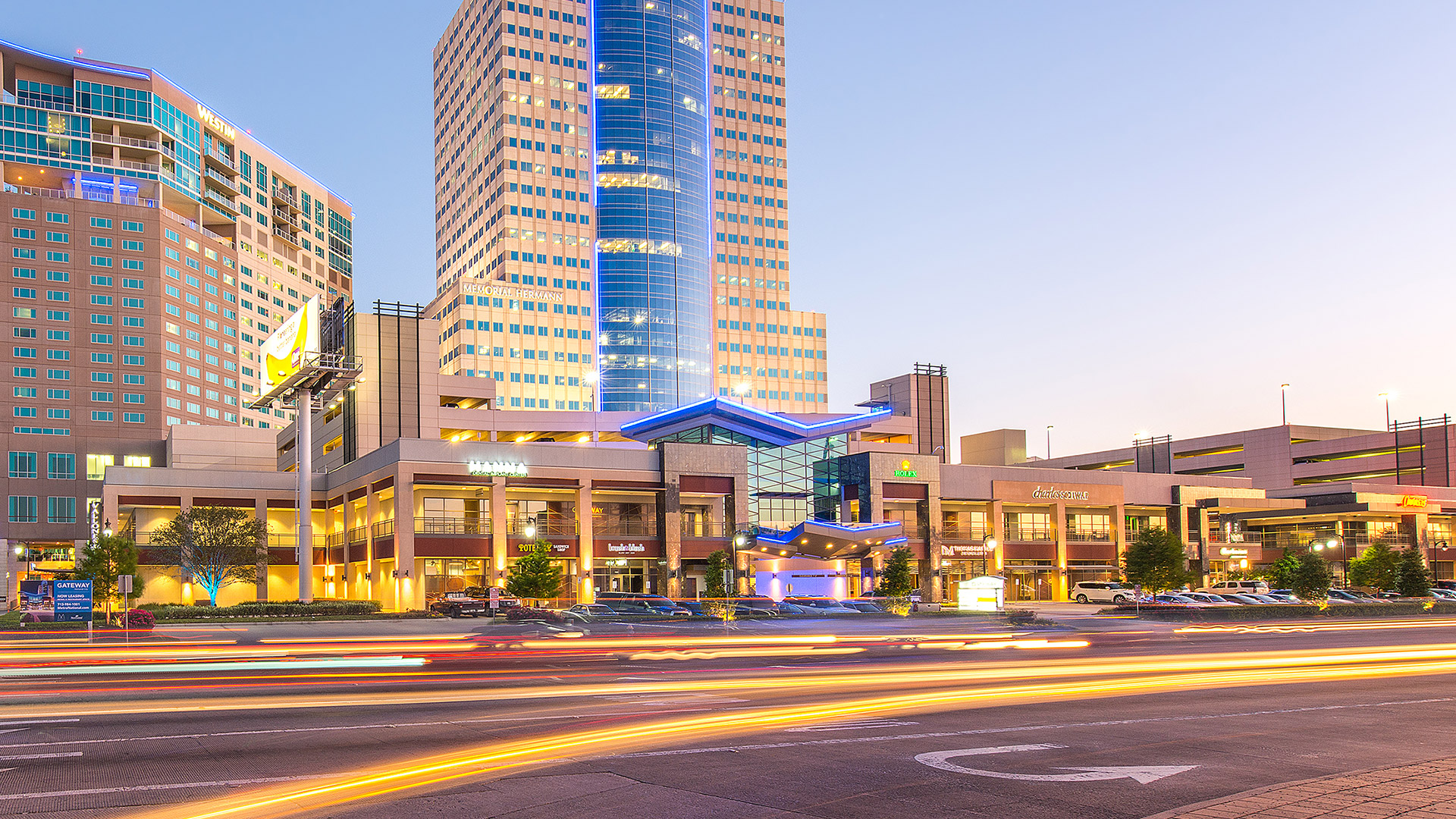 Street view of Memorial City at dusk, with lit buildings and traffic light trails