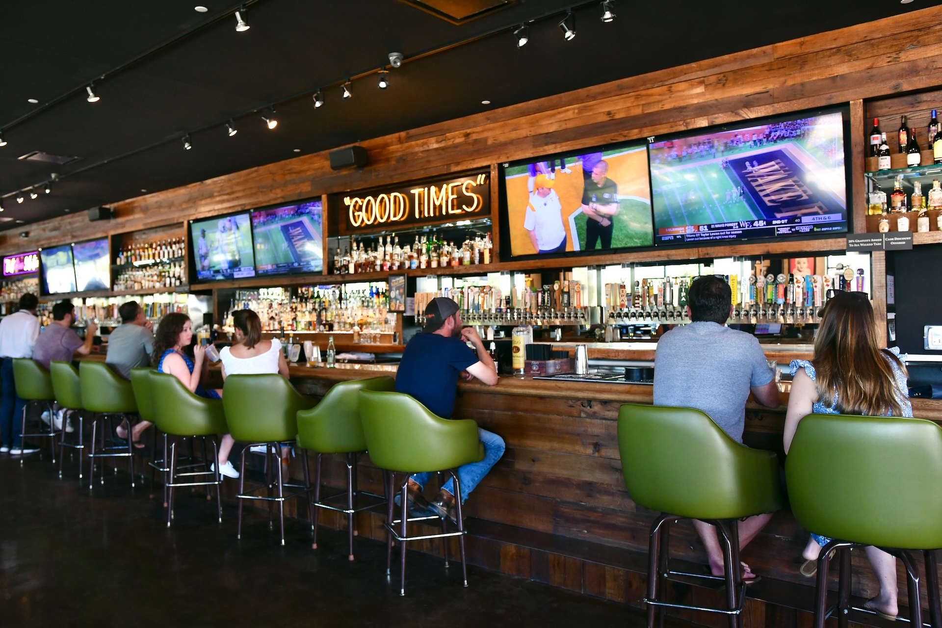 Patrons seated at the bar of a sports bar at Greenside, with green stools and a wall of television screens
