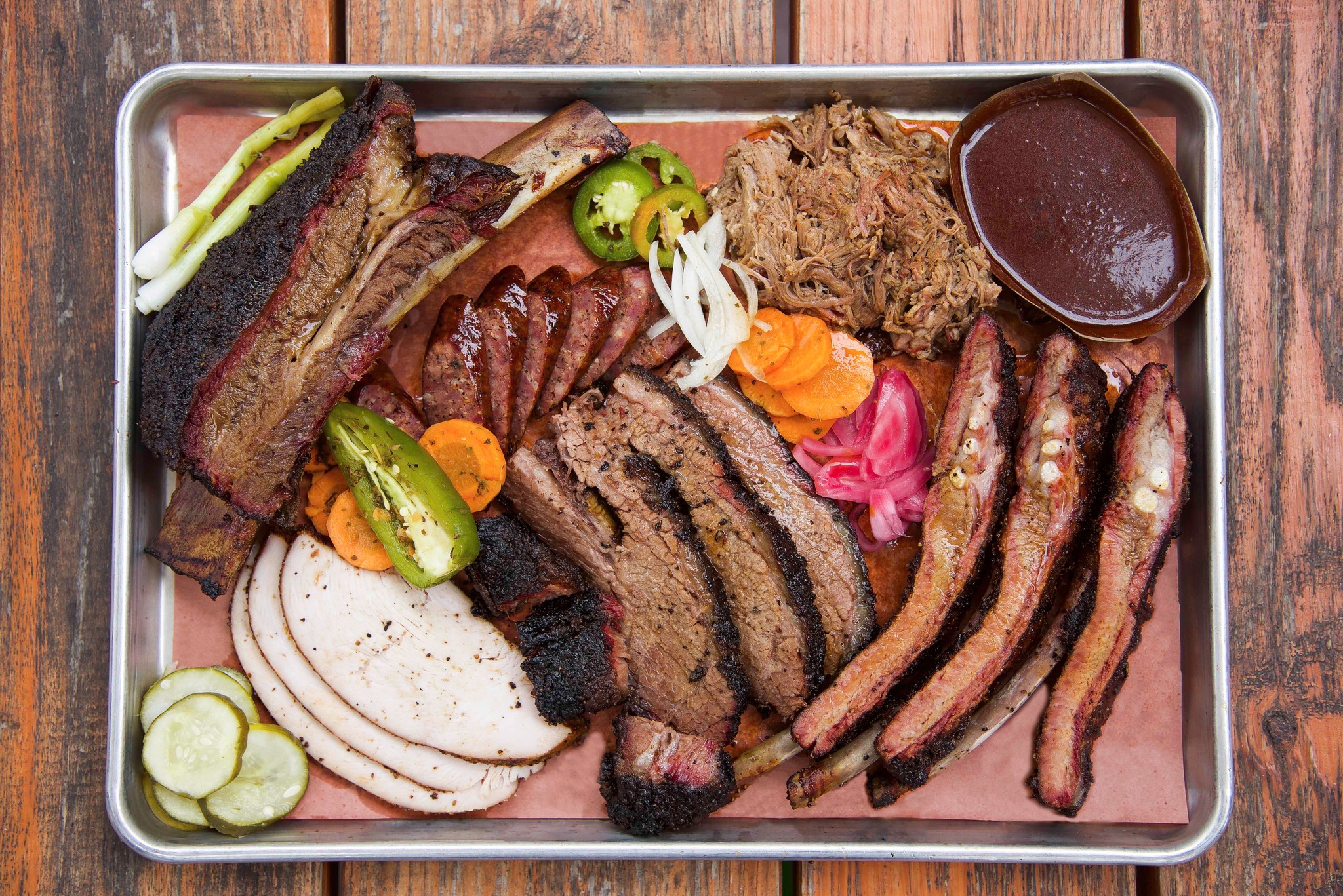 Metal tray of barbecue with sliced brisket, sausage links, turkey, pickles, onions and a cup of sauce