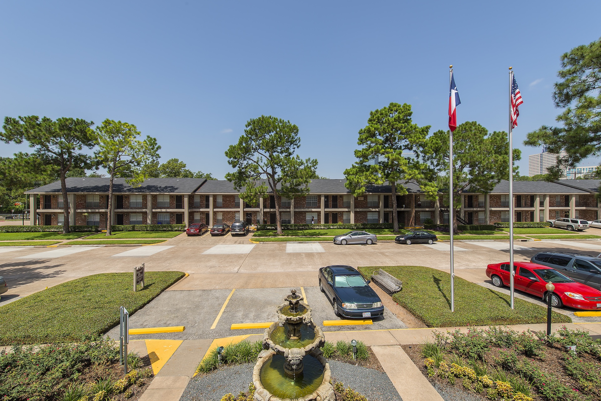 Exterior of Memorial City Apartments with a fountain, flagpoles and mature shade trees along the parking area