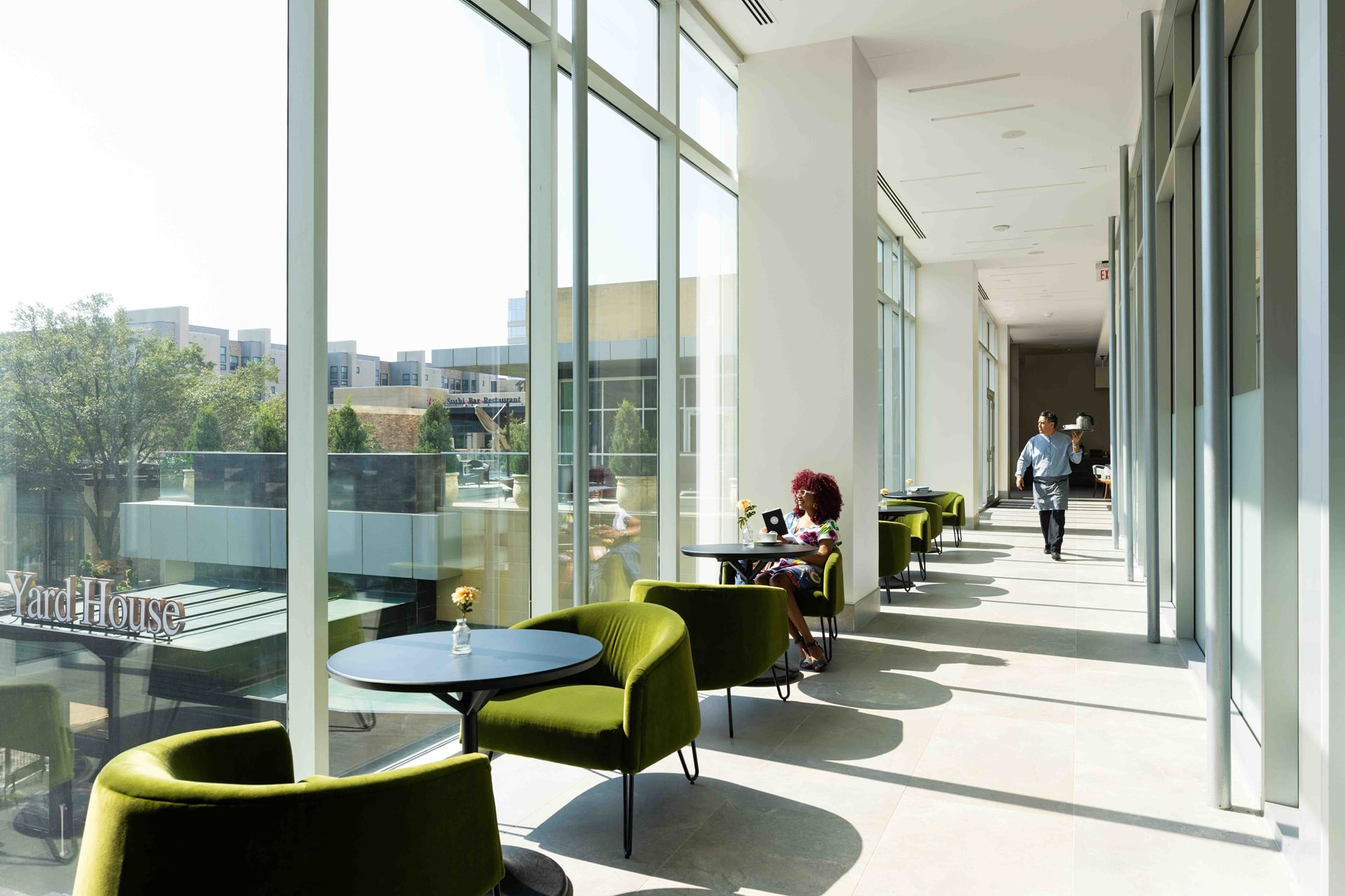 Glass-walled lounge corridor at The Moran Hotel with green armchairs and cafe tables