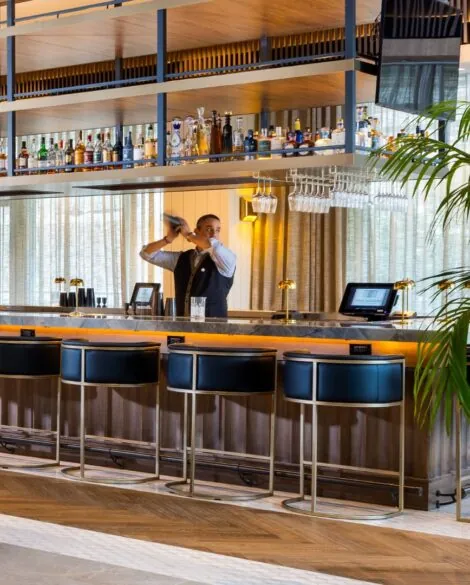 Bartender behind the bar at The Moran Hotel, with blue bar stools and a lit bottle display